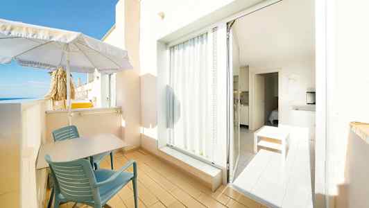 En balkong med bord och stolar, parasoll och utsikt mot havet. En dörr leder till ett ljus interiör med möbler. (AI-genererad text)