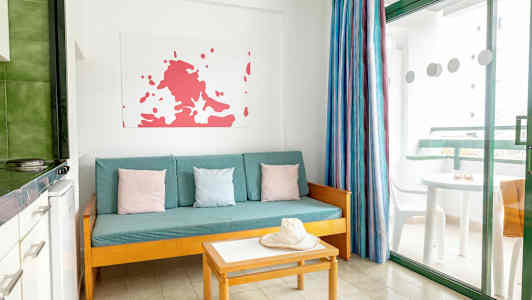 Ett vardagsrum med en turkos soffa, färgglada kuddar, bord och en hatt. Ett fönster med utsikt mot en balkong. (AI-genererad text)