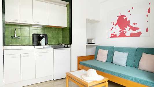 Ett enkelt kök med vit inredning och gröna kakel bakom. Brevid finns en soffa med turkosa dynor och pastellfärgade kuddar. (AI-genererad text)