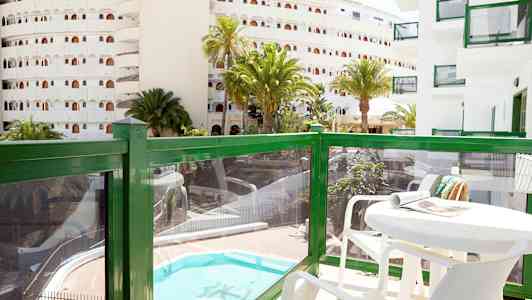 Balkong med bord och stolar, utsikt över en pool och omgivande byggnader. (AI-genererad text)