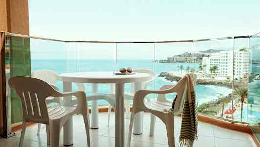 Utsikt från en balkong med ett bord och plaststolar, havet och byggnader synliga i bakgrunden. (AI-genererad text)