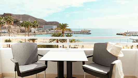 Två stolar och ett bord på en balkong med utsikt över en strand och havet. (AI-genererad text)