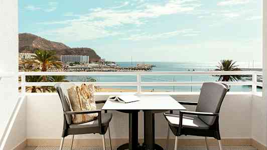 Utsikt från en balkong med bord och stolar mot en strand och havet. (AI-genererad text)