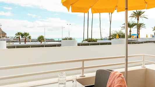 En balkong med ett bord, en orange handduk på en stol, en karaff och ett glas på bordet samt gula parasoller och palmer i bakgrunden. (AI-genererad text)