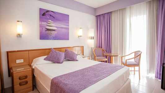 Hotellrum med dubbelsäng, lila detaljer och sittplats vid ett fönster. (AI-genererad text)