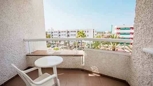 Bild av en balkong med ett bord och en stol, med utsikt över byggnader och palmträd. (AI-genererad text)