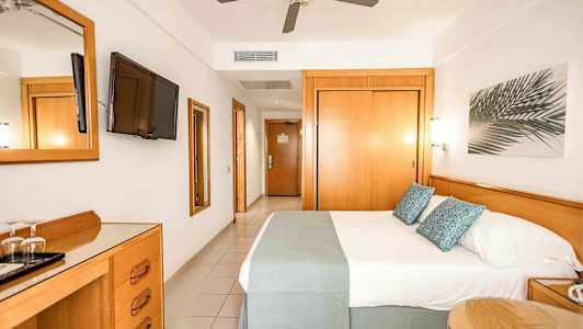 Ett hotellrum med en dubbelsäng, TV på väggen, takfläkt och möbler i trä. (AI-genererad text)