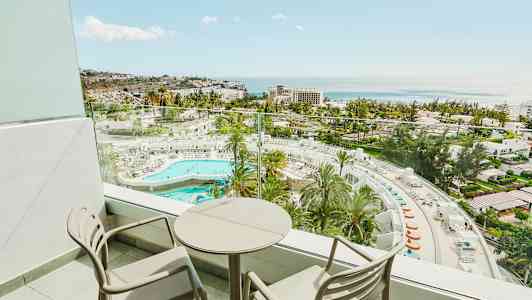 Balkong med två stolar och ett runt bord samt utsikt över hotellområde med pool och hav. (AI-genererad text)