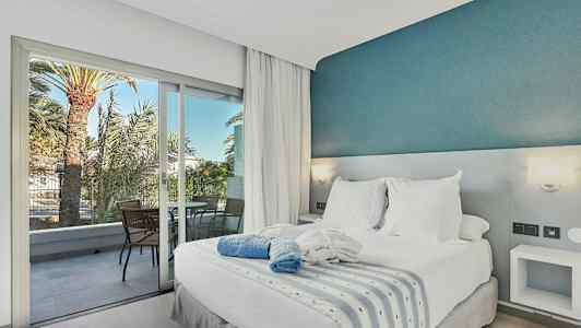 Sovrum med dubbelsäng, vitt sänglinne och blå dekorativ vägg, samt balkong med stolar och utsikt över palmer. (AI-genererad text)