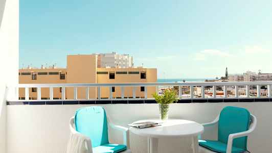 En balkong med två stolar, ett bord och utsikt över byggnader och havet. (AI-genererad text)