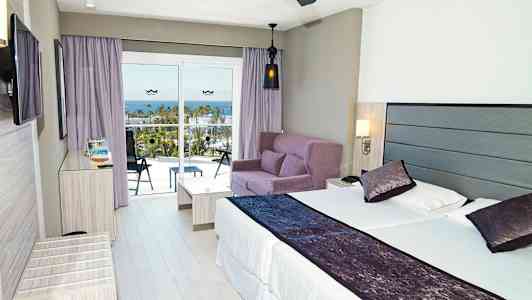 Modernt hotellrum med en dubbelsäng, soffgrupp och balkong med utsikt över havet. (AI-genererad text)