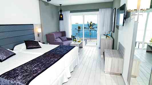 Hotellrum med dubbelsäng, soffa och havsutsikt utanför stora fönster. (AI-genererad text)