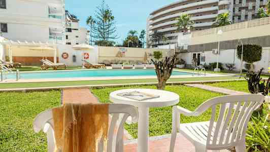 En utomhuspool omgiven av grönytor och solstolar, med vita byggnader i bakgrunden. (AI-genererad text)