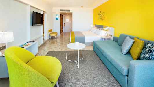 Modernt möblerat hotellrum med färgglada detaljer som gul stol och blå soffa. (AI-genererad text)