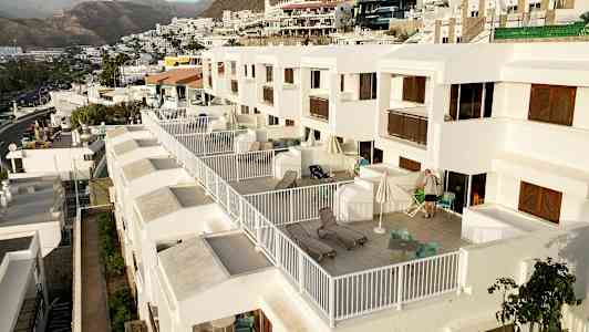 Bild på en rad av moderna vita lägenheter med stora privata balkonger, möblerade med stolar och solstolar, i en kuperad miljö med berg i bakgrunden. (AI-genererad text)