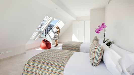 Ett ljust rum med två sängar, färgglada dynor, en röd sittpuff och ett stort lutande fönster. (AI-genererad text)