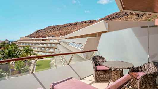 En terrass med solstolar och möbler, med utsikt över hotellbyggnader, palmträd och berg. (AI-genererad text)