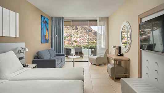 Hotellrum med två sängar, soffa, fåtölj och stor balkong med utsikt över en bergig stadsdel. (AI-genererad text)