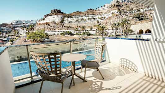 En balkong med två stolar och ett bord, utsikt över en pool, byggnader och berg. (AI-genererad text)