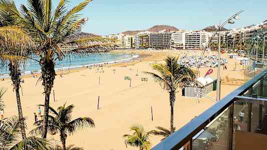 Vy över en bred sandstrand med palmer och stadens byggnader i bakgrunden. (AI-genererad text)