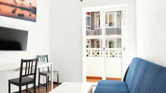 Interiörbild av vardagsrum med en blå soffa, ett vitt bord, två svarta stolar och ett TV på väggen. Bakom glasdörren syns en byggnad. (AI-genererad text)