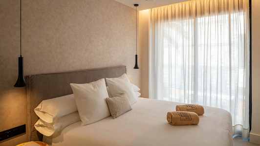 Ett hotellrum med en stor säng, vita lakan och kuddar. På sängen ligger hoprullade handdukar. Väggar och gardiner är ljusa, och rummet har hängande lampor. (AI-genererad text)