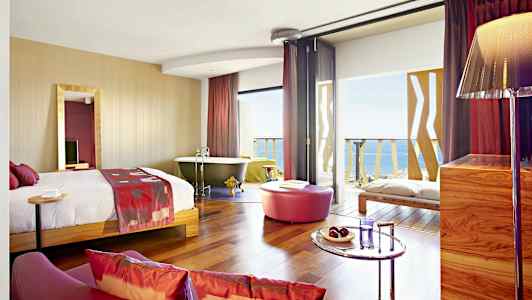 Ett hotellrum med en säng, badkar, sittgrupp och utsikt över havet. (AI-genererad text)