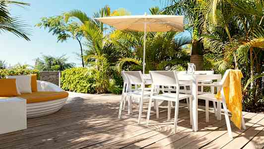 En uteplats med vita stolar och bord under ett parasoll, omgiven av gröna växter. (AI-genererad text)