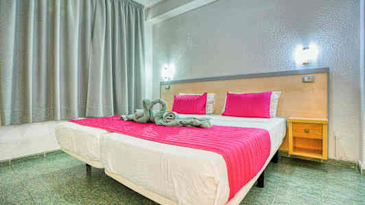 Säng med rosa täcke och kuddar i ett rum med grå gardiner och en träbänk. (AI-genererad text)