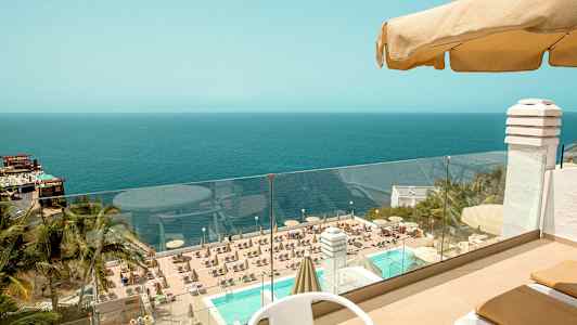 Utsikt från en terrass med bord och stolar ut mot en pool omgiven av solsängar och parasoller, med en vy över havet i bakgrunden. (AI-genererad text)