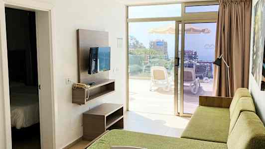 En hotellrumsvy med en grön soffa och en TV på väggen, med en dörröppning till ett sovrum och en terrass som visar omgivningen utanför. (AI-genererad text)