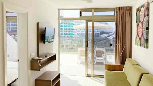 Ett ljust hotellrum med soffa, väggmonterad TV och glidande glasdörr som leder ut till en möblerad balkong. (AI-genererad text)