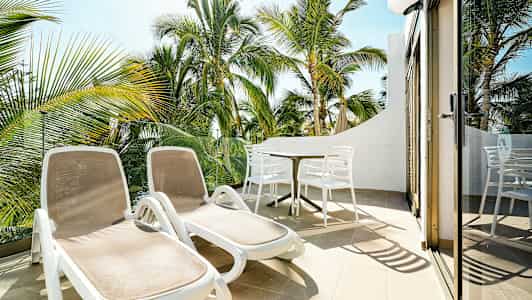 Solstolar och matbord på en terrass med utsikt över palmträd. (AI-genererad text)