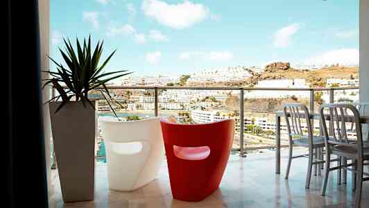 En balkong med utsikt över en stad och berg, med två moderna stolar och en växt i kruka. (AI-genererad text)