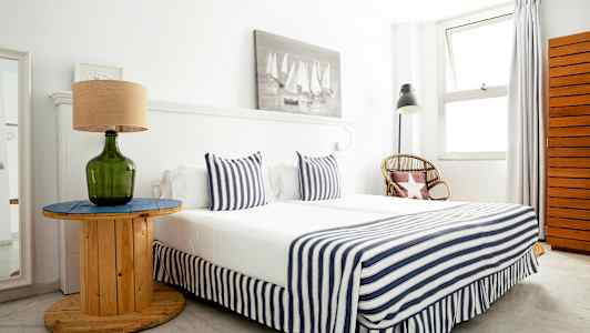 Ett sovrum med en säng klädd i randiga textilier, ett bord med en grön glaslampa och en rottingstol vid sidan. (AI-genererad text)