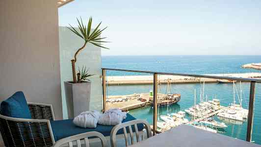 Utsikt över en hamn med flera båtar från en balkong med stolar och en krukväxt. (AI-genererad text)