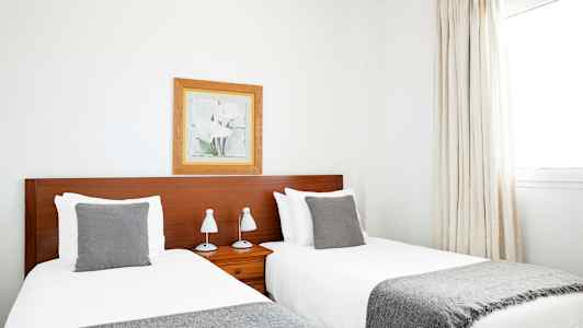 Två enkelsängar med gråa filtar och kuddar, placerade bredvid en träfärgad huvudgavel. Ett inramat porträtt hänger på väggen ovanför huvudgaveln. (AI-genererad text)