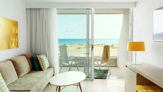 Utsikt från ett ljust hotellrum med modern inredning över en solig strand och havet. (AI-genererad text)