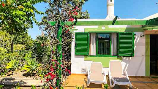 Bild av ett hus med gröna fönsterluckor och en uteplats med två solstolar. Planteringar med blommor och växter omger området. (AI-genererad text)