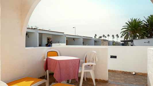 Den visar en uteplats med solstolar och ett bord med stolar. Bakgrunden har små hus och palmer. (AI-genererad text)