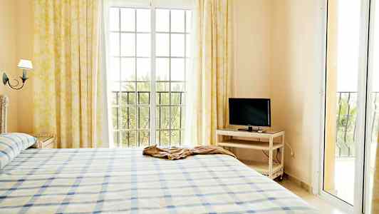 En ljus sovrumsinteriör med en säng, en tv på ett bord och stora fönster med gardiner. (AI-genererad text)