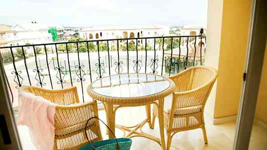 Två rottingstolar och ett runt bord med glasskiva står på en balkong med utsikt över kortstensbyggnader och ett tennisområde. (AI-genererad text)