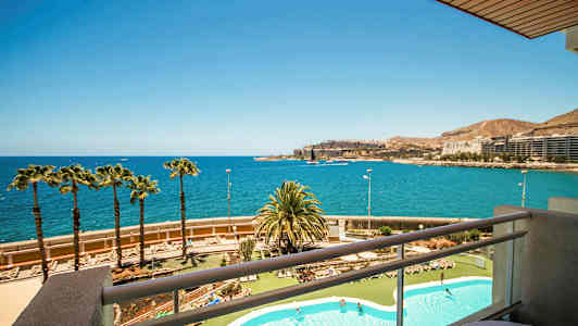 Utsikt från en balkong över ett poolområde och en kustlinje med palmer, havet och byggnader i fjärran. (AI-genererad text)