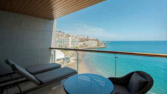 Utsikt från en balkong över havet och en strand, med bord och stolar samt solstolar i förgrunden. (AI-genererad text)