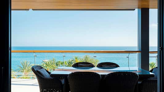 Bild av en terrass med bord och stolar, havsutsikt och palmer i bakgrunden. (AI-genererad text)