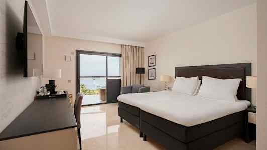 Ett hotellrum med en stor säng, ett skrivbord och en TV. Genom glasdörren syns en balkong och utsikt mot havet. (AI-genererad text)