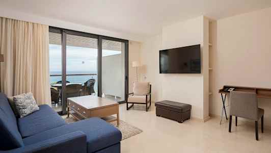 Ett vardagsrum med blå soffa, träbord och stolar vid ett stort fönster mot balkongen. (AI-genererad text)