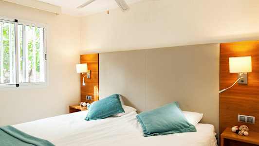 Ett ljust sovrum med en dubbelsäng, blå kuddar, och fönster. (AI-genererad text)