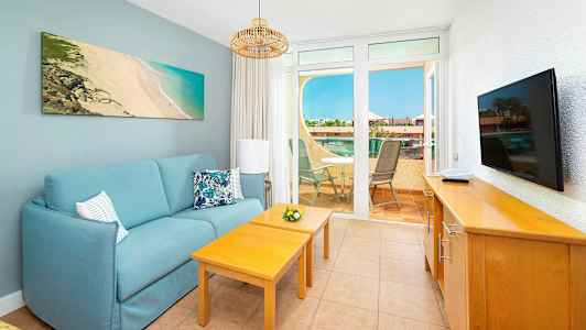 Ett vardagsrum med en blå soffa, två träbord, en TV på en byrå och utsikt över en terrass med utemöbler. (AI-genererad text)