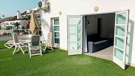 Utomhusområde med grön gräsmatta, sittgrupp och öppna dörrar till en interiör. (AI-genererad text)